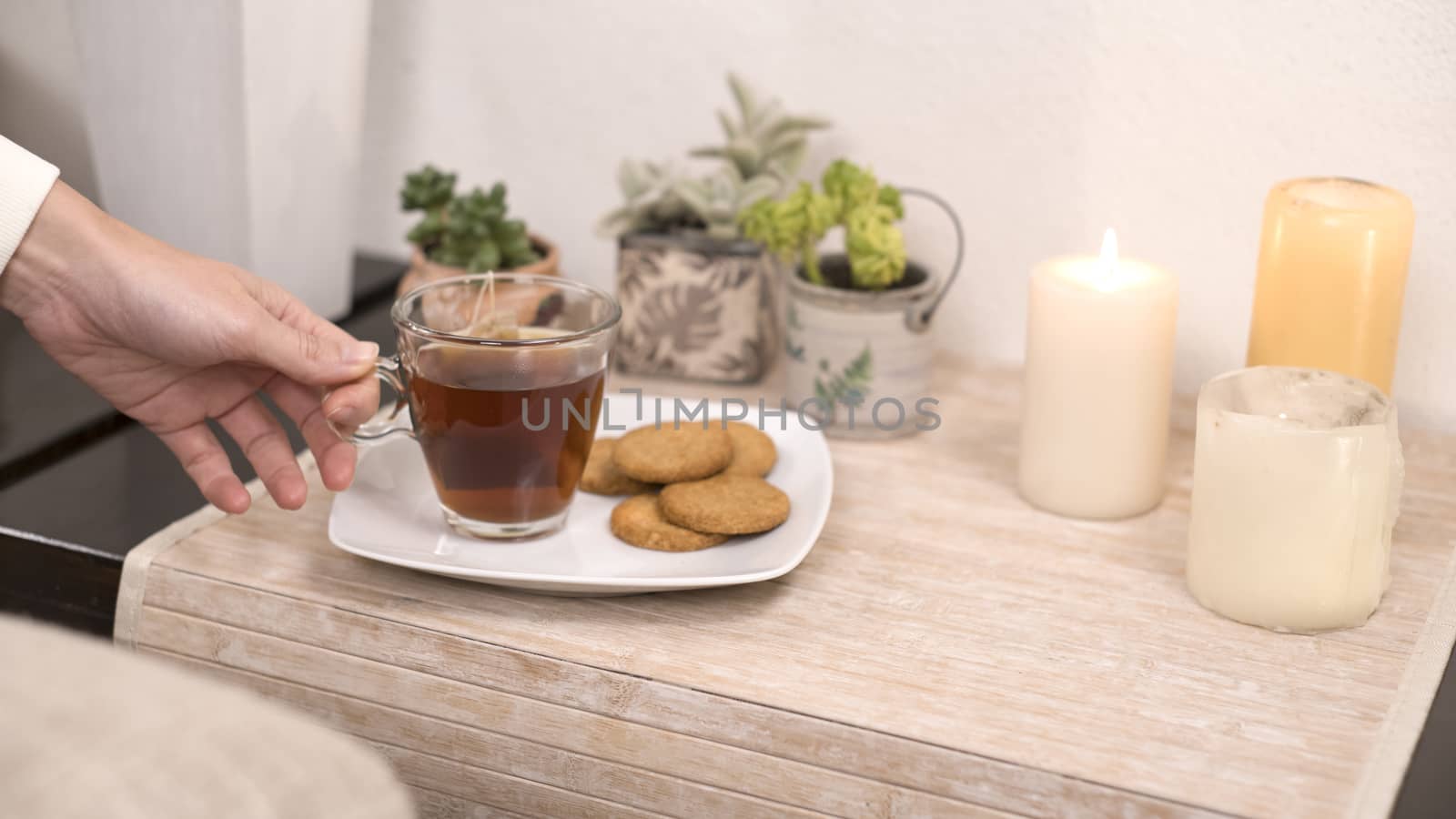 Relaxation concept: woman's hand takes a cup of tea from a plate with biscuits on a small table with candles little plants in bokeh effect