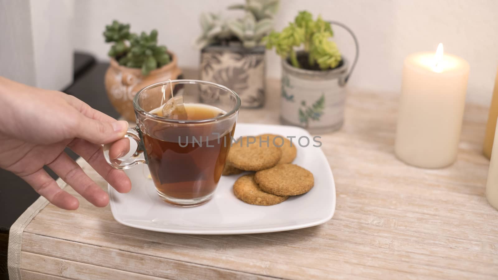 Relaxation concept: woman's hand takes a cup of tea from a plate with biscuits on a small table with candles little plants in bokeh effect