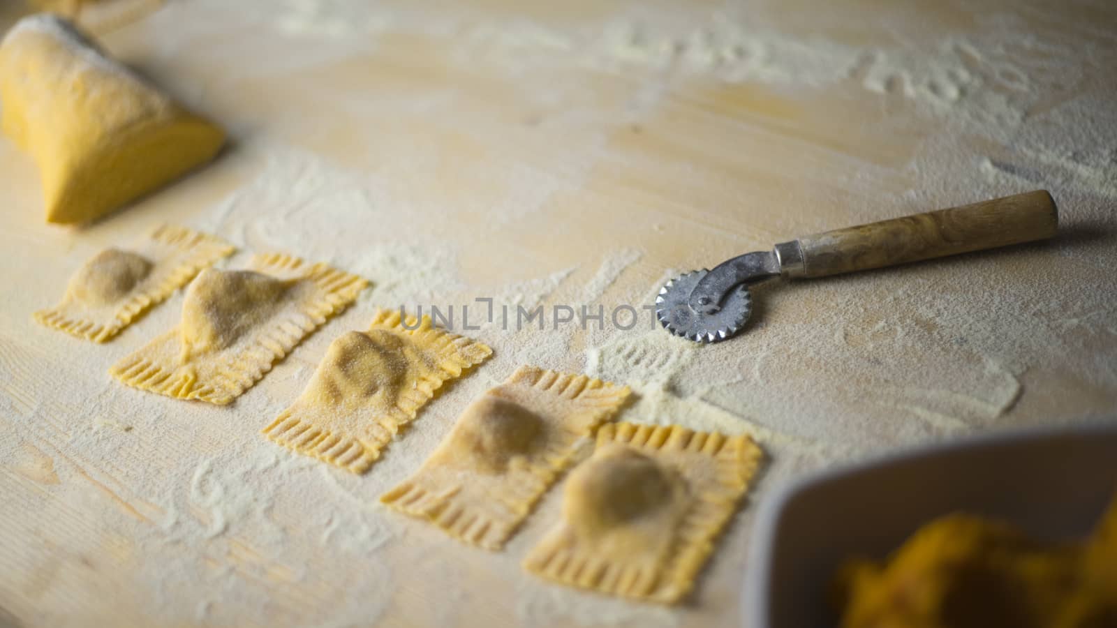 Closeup of pumpkin ravioli vegan, traditional Italian pasta, homemade pasta just made, with the dough mass durum wheat flour, the cutting wheel and wheat flour scattered on the light wooden table by robbyfontanesi