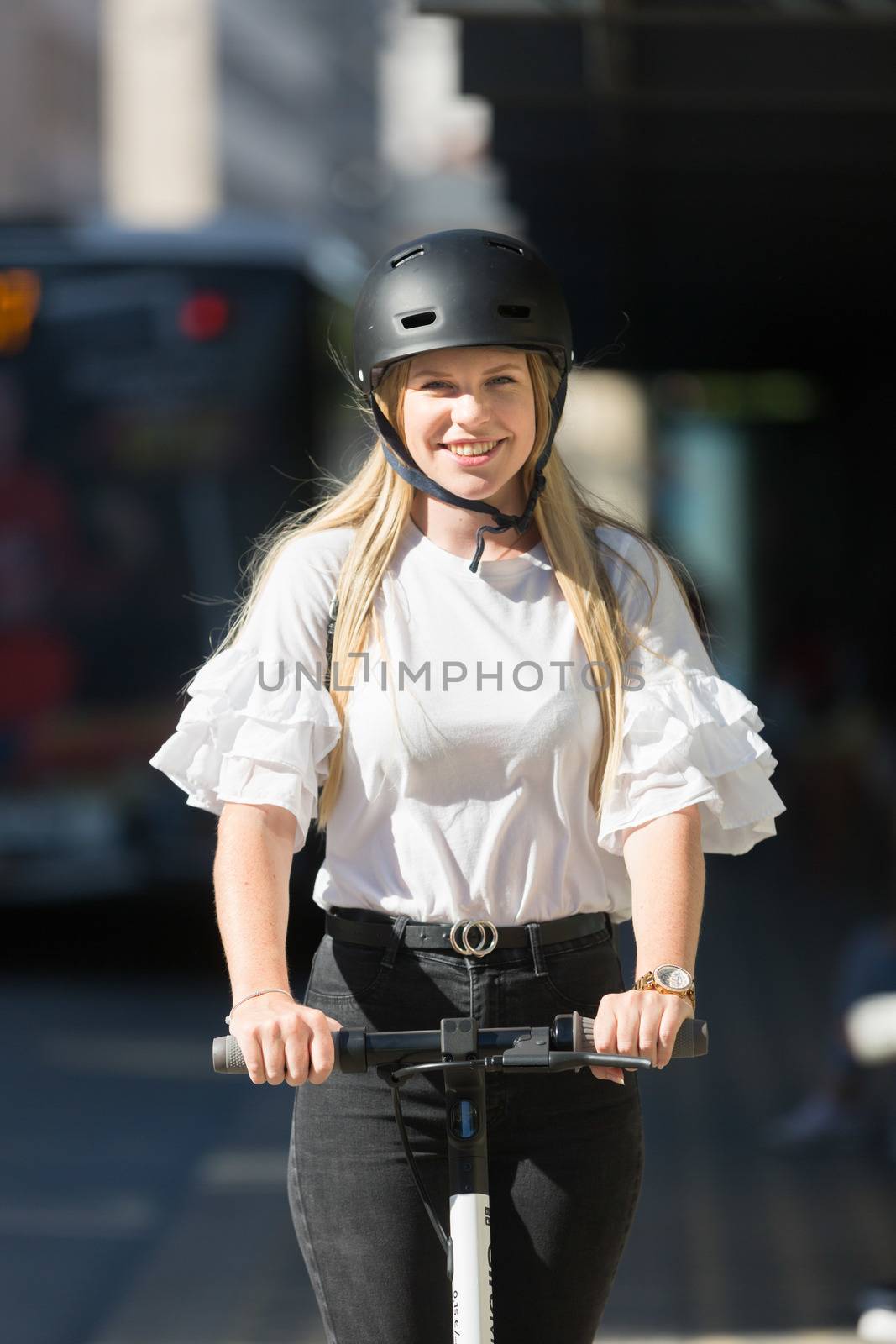Trendy fashinable teenager, beautiful blonde girl riding public rental electric scooter in urban city environment. Eco-friendly modern public city transport.