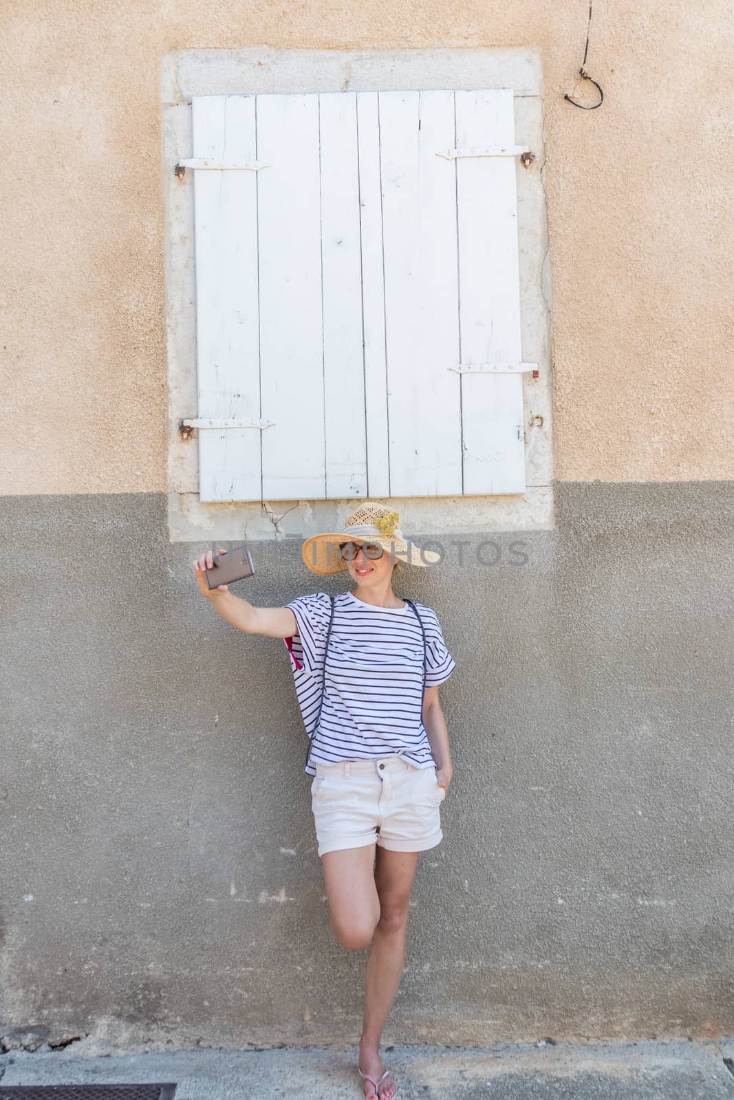Beautiful young female tourist woman wearing big straw hat, taking self portrait selfie, standing in front of white vinatage wooden window and textured stone wall at old Mediterranean town by kasto