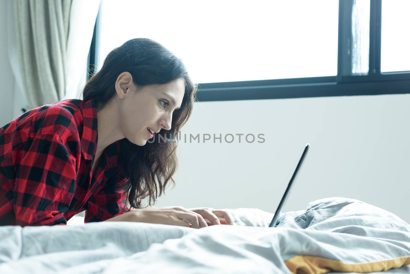 Beautiful woman working on a laptop with smiling and lying down on the bed at a condominium in the morning.