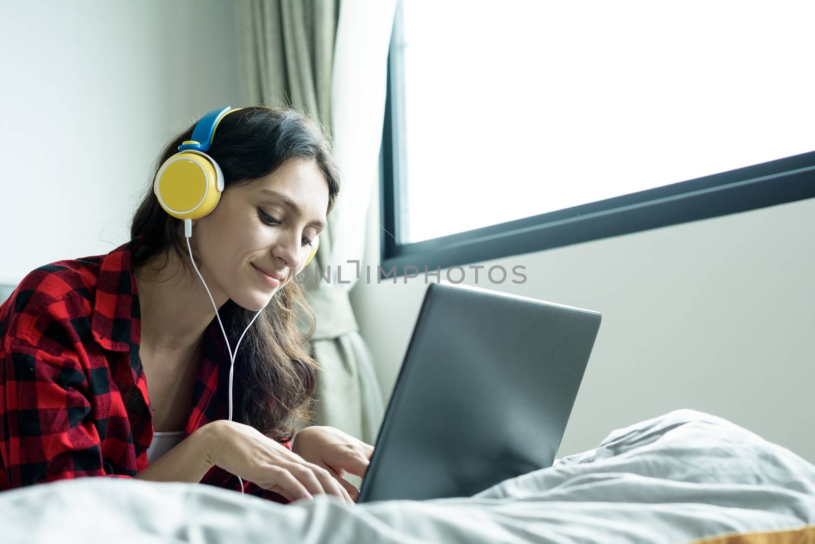 Beautiful woman working and listening to music on a laptop with yellow headphone and lying down on the bed at a condominium in the morning.