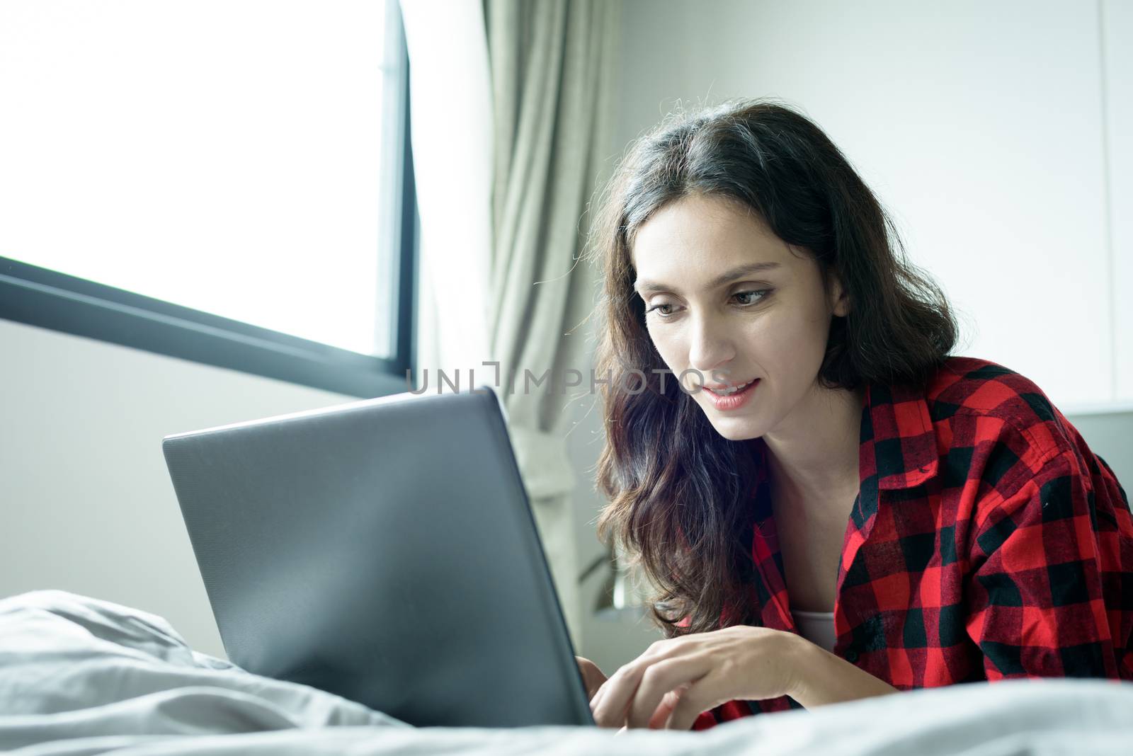 Beautiful woman working on a laptop with smiling and lying down on the bed at a condominium in the morning.