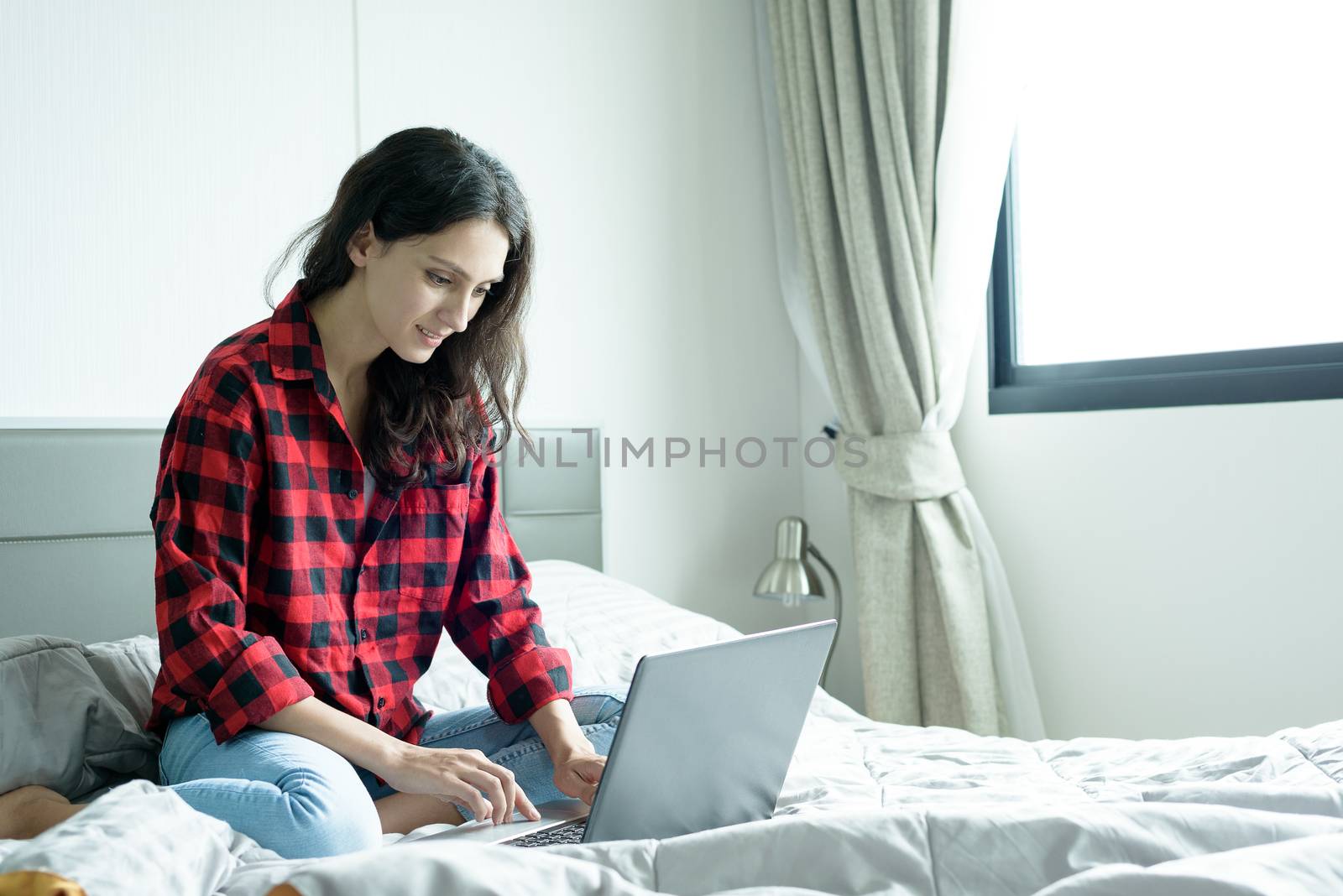 Beautiful woman working on a laptop with smiling and sitting on the bed at a condominium in the morning.