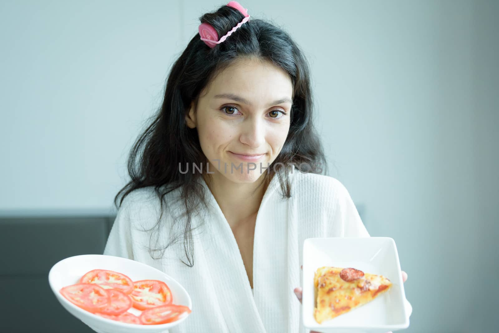 A beautiful woman wearing a towel and a white bathrobe has to eat a pizza and sliced tomato with happy and relaxing on the bed at a condominium in the morning.