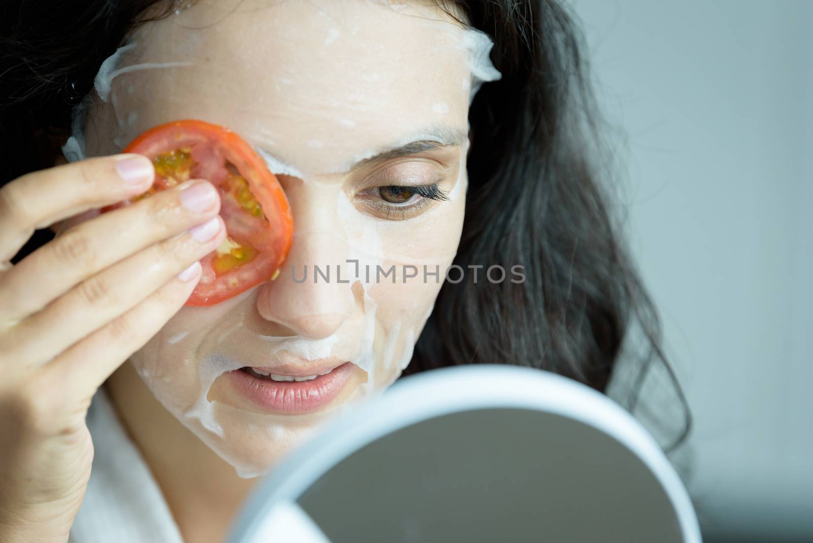 A beautiful woman wearing a towel and a white bathrobe has to look to a mirror for a mask on face-covering eyes with sliced tomatoes with happy and relaxing on the bed at a condominium in the morning.