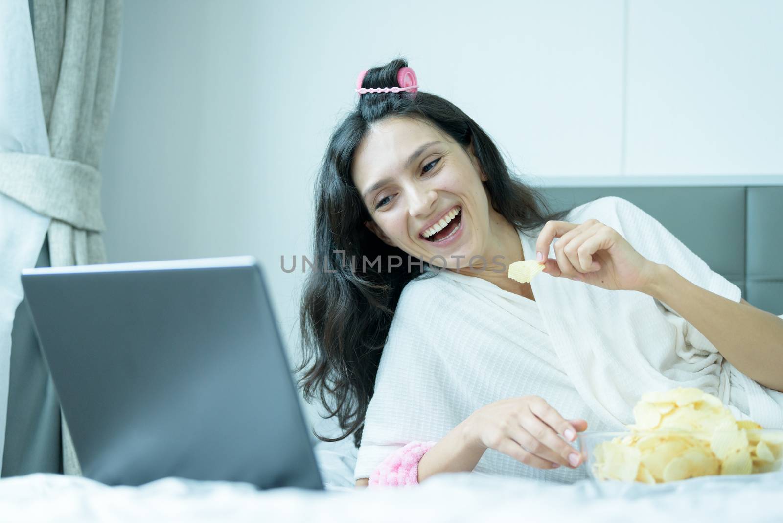 A beautiful woman eating snack potato and playing laptop with lying down on the bed and happiness at a condominium in the morning.