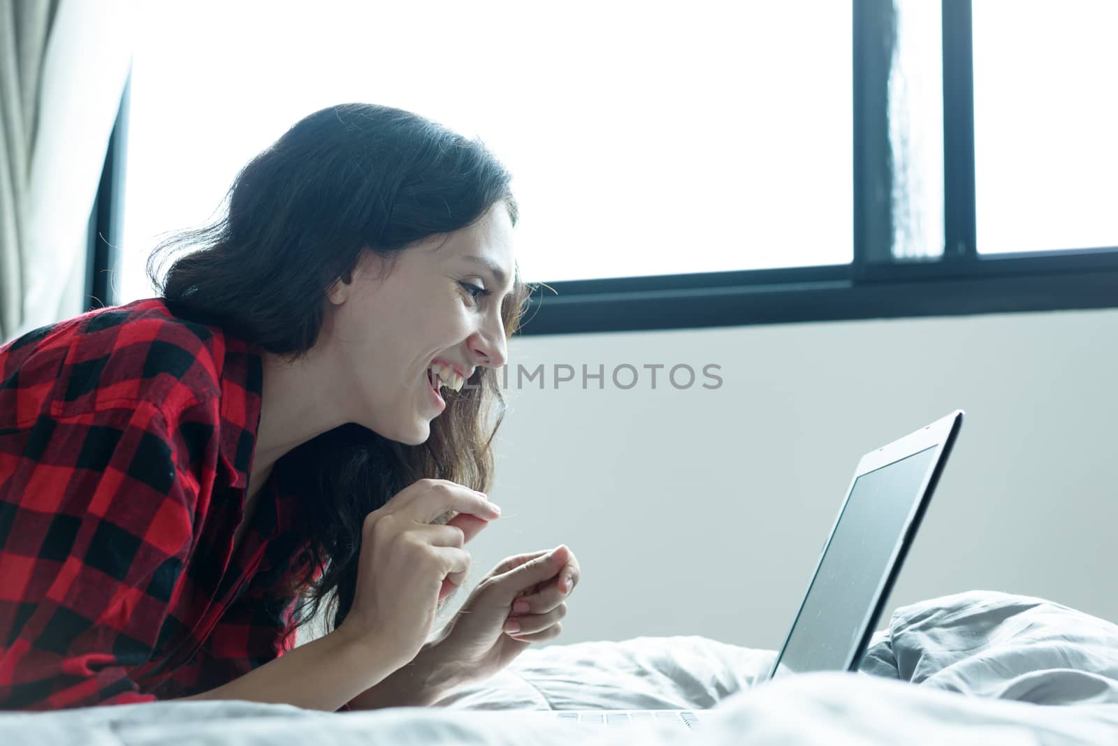 Beautiful woman working on a laptop with smiling and lying down on the bed at a condominium in the morning.