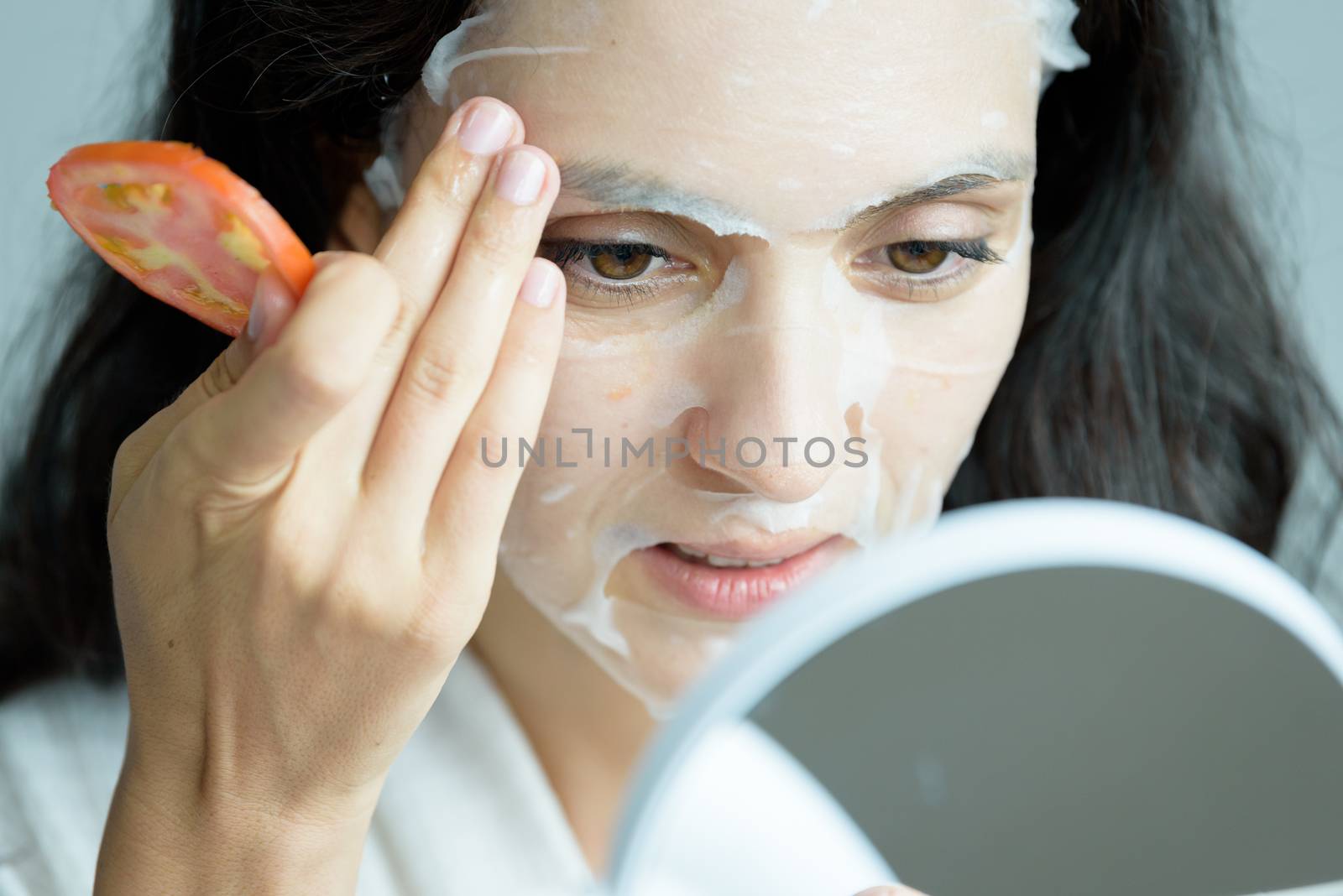 A beautiful woman wearing a towel and a white bathrobe has to look to a mirror for a mask on face-covering eyes with sliced tomatoes with happy and relaxing on the bed at a condominium in the morning.