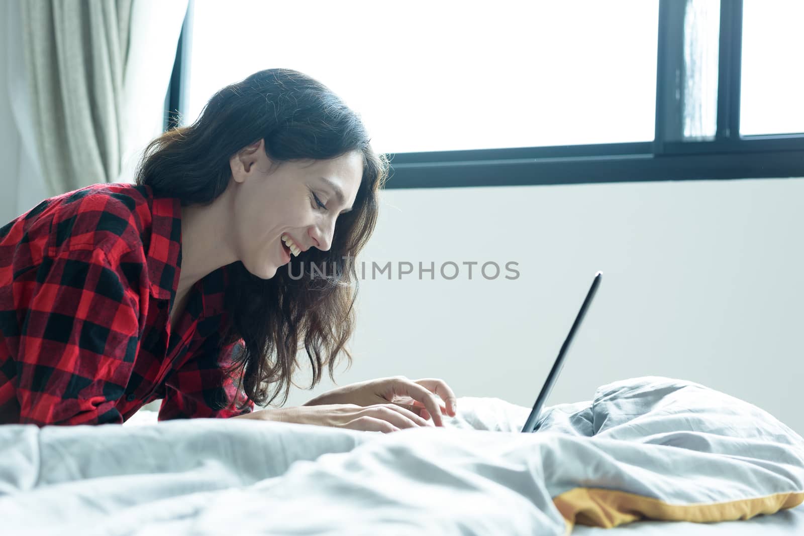 Beautiful woman working on a laptop with smiling and lying down on the bed at a condominium in the morning.