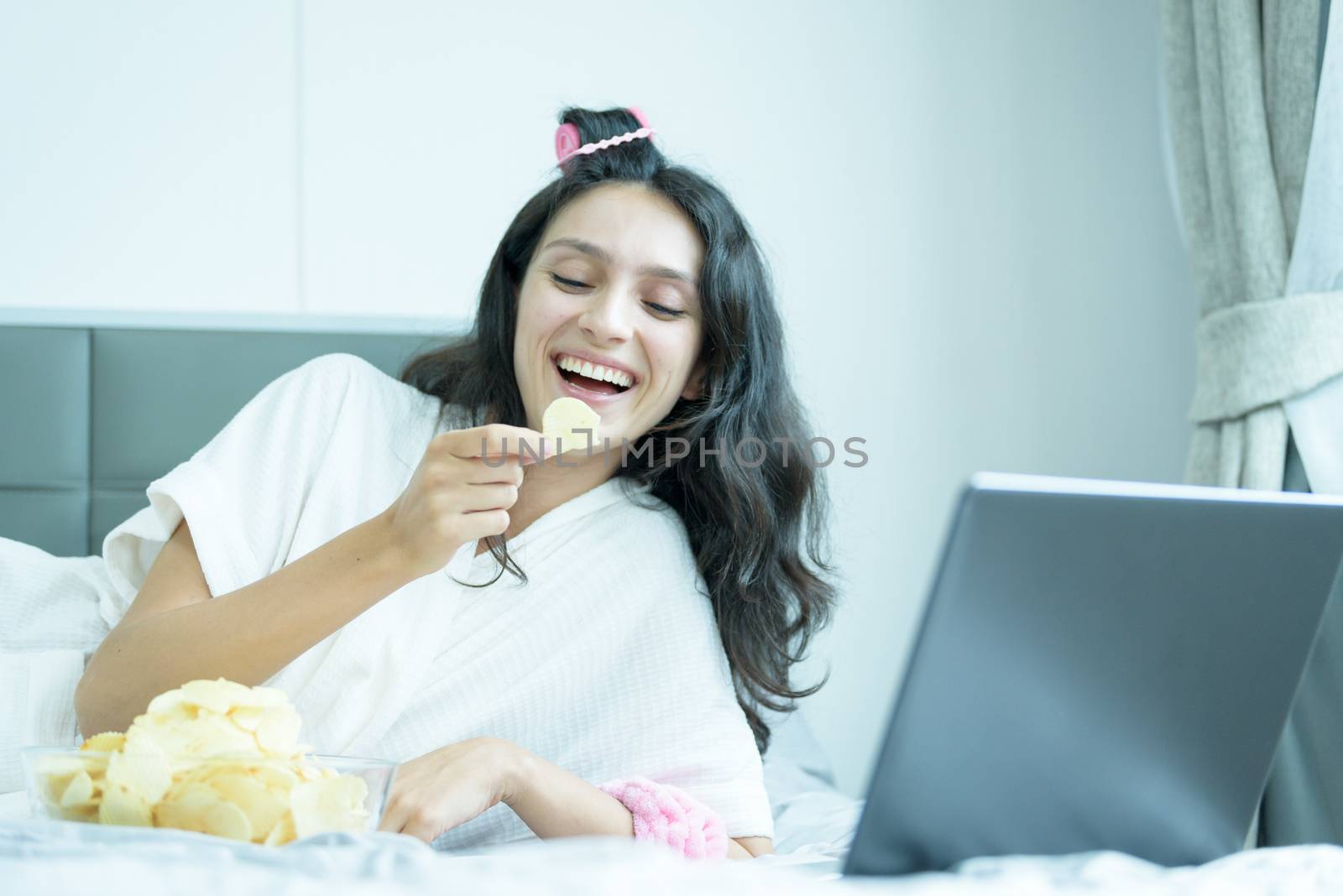 A beautiful woman eating snack potato and playing laptop with lying down on the bed and happiness at a condominium in the morning.