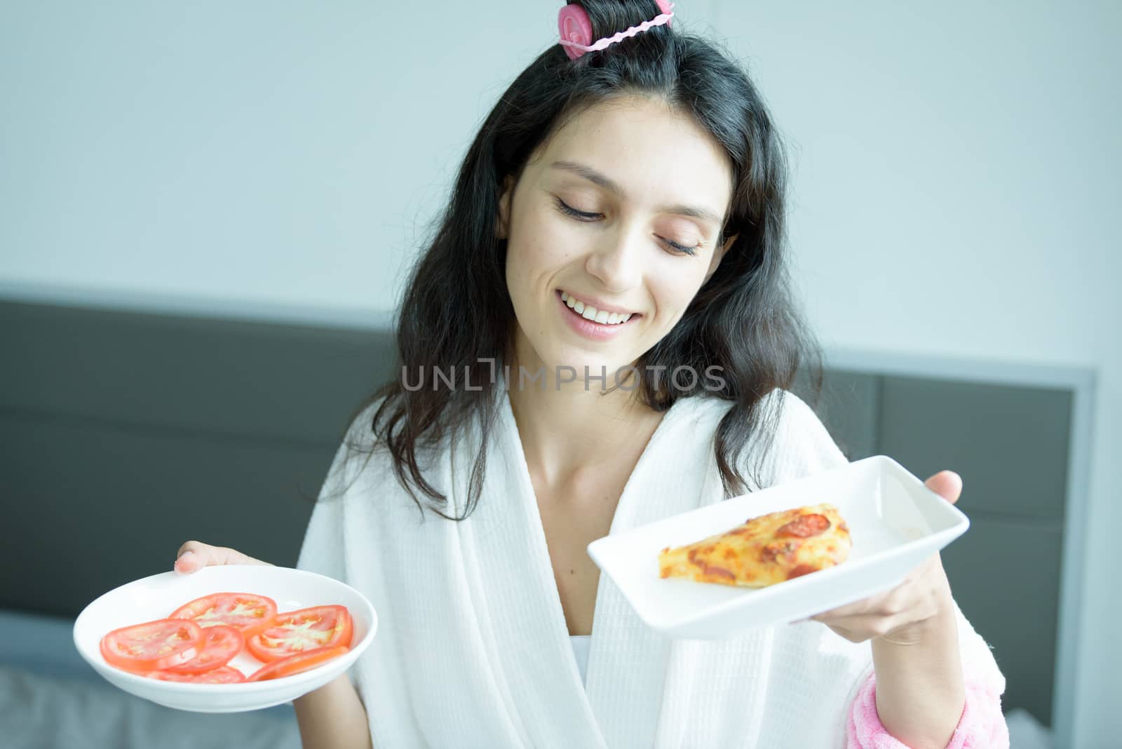 A beautiful woman wearing a towel and a white bathrobe has to eat a pizza and sliced tomato with happy and relaxing on the bed at a condominium in the morning.
