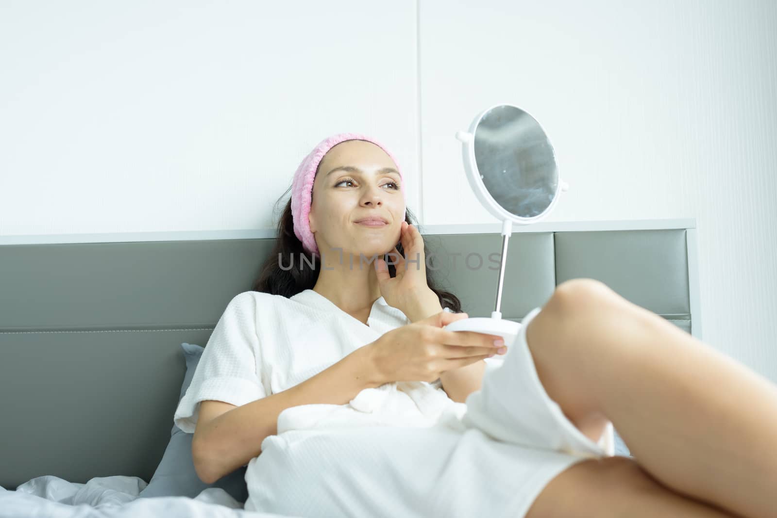 A beautiful woman wearing a towel and a white bathrobe and pink headband has to look a mirror with happy and relaxing on the bed at a condominium in the morning.