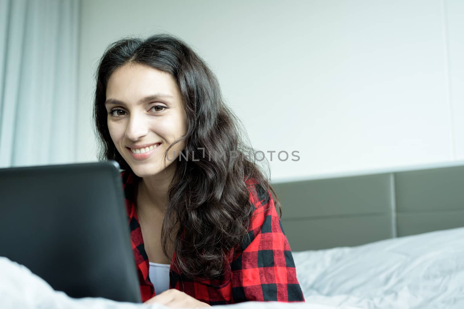 Beautiful woman working on a laptop with smiling and lying down on the bed at a condominium in the morning.