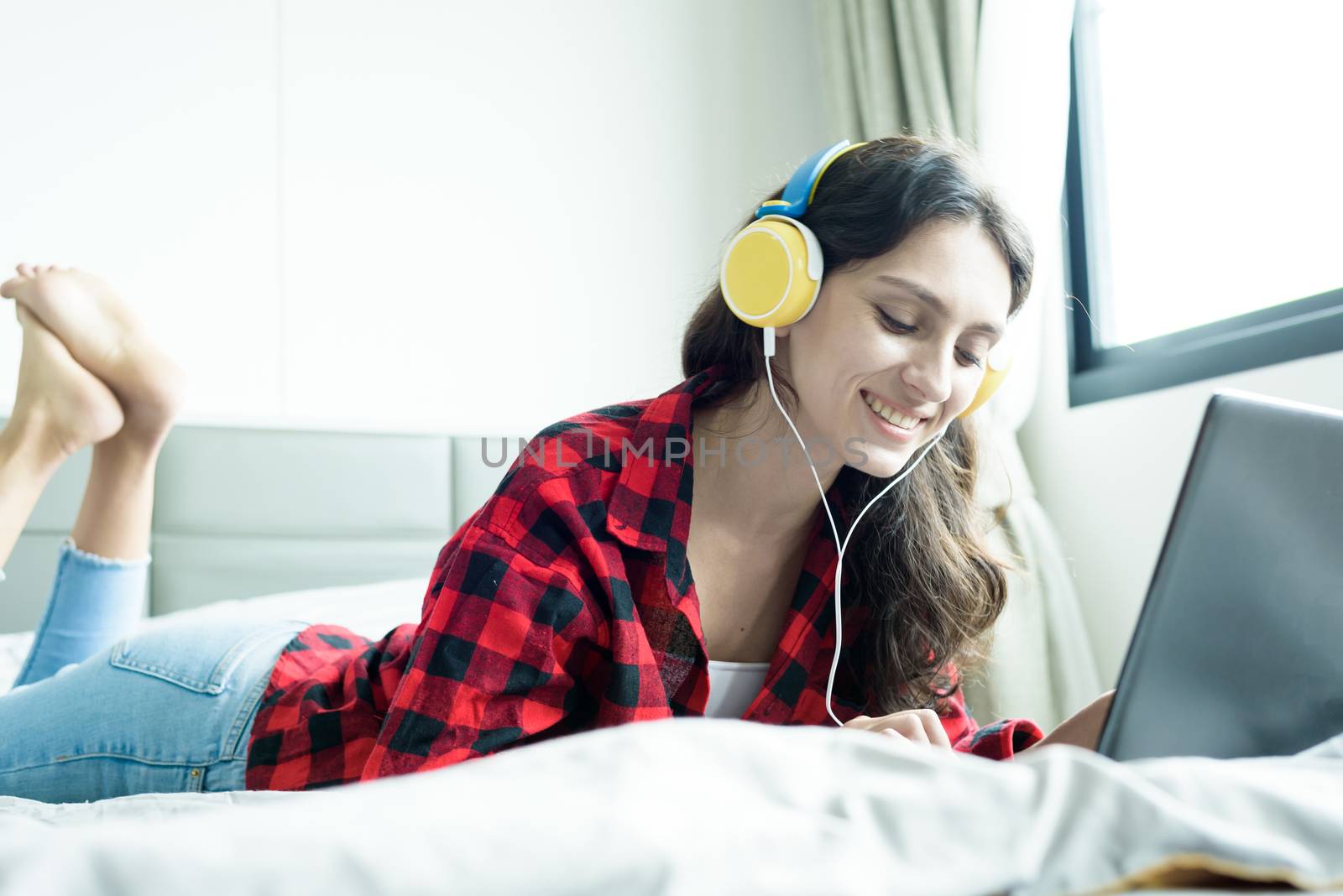 Beautiful woman working and listening to music on a laptop with yellow headphone and lying down on the bed at a condominium in the morning.