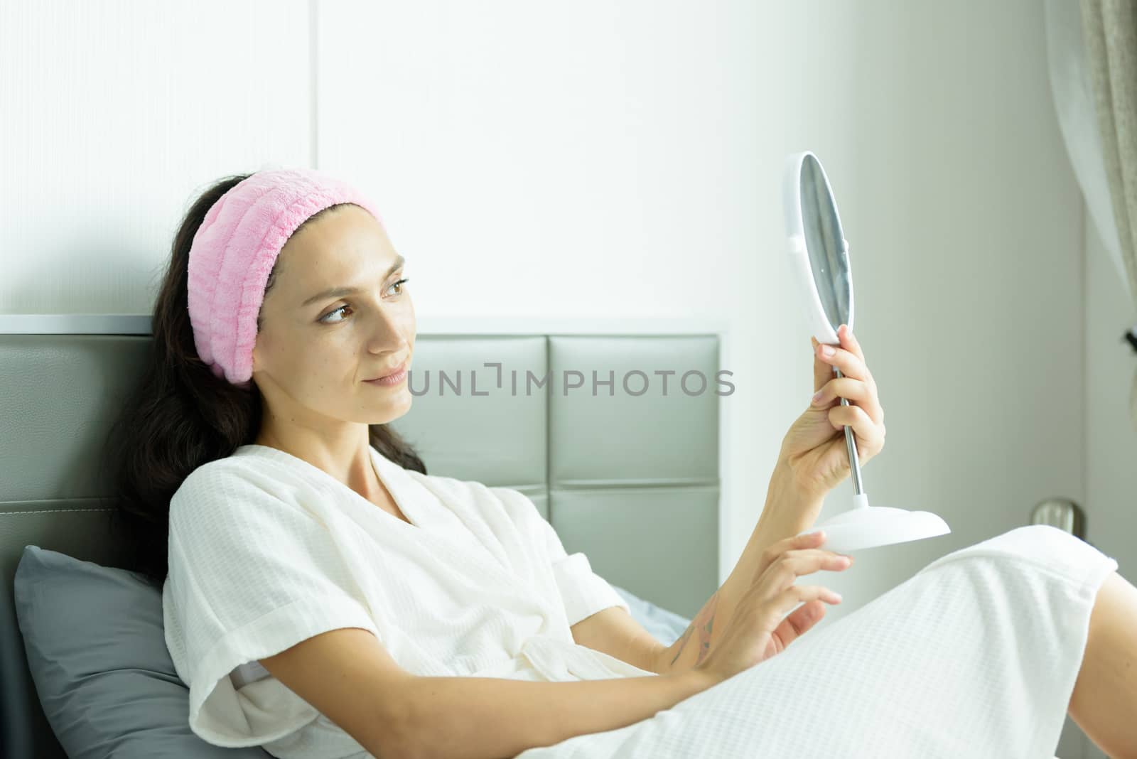 A beautiful woman wearing a towel and a white bathrobe and pink headband has to look a mirror with happy and relaxing on the bed at a condominium in the morning.