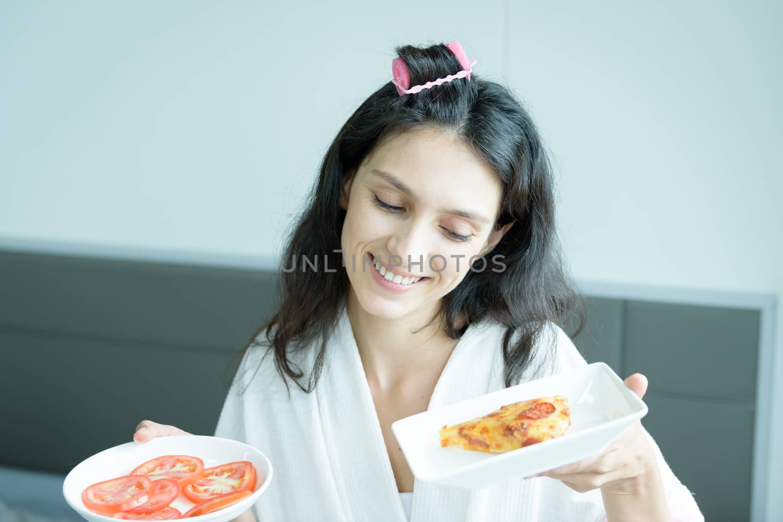 A beautiful woman wearing a towel and a white bathrobe has to eat a pizza and sliced tomato with happy and relaxing on the bed at a condominium in the morning.