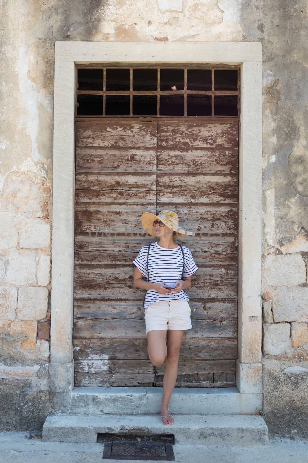 Beautiful young female tourist woman standing in front of vinatage wooden door and textured stone wall at old Mediterranean town, smiling, holding, smart phone to network on vacationes by kasto