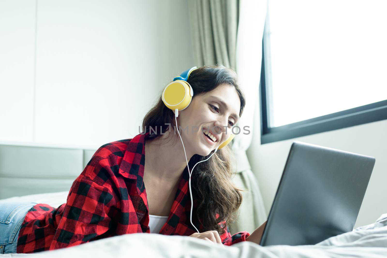 Beautiful woman working and listening to music on a laptop with yellow headphone and lying down on the bed at a condominium in the morning.