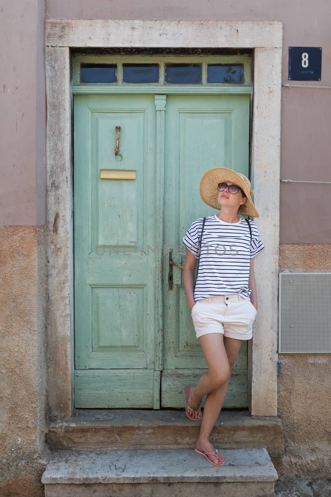 Beautiful young female tourist woman wearing sun hat, standing and relaxing in shade in front of vinatage wooden door in old Mediterranean town while sightseeing on hot summer day by kasto
