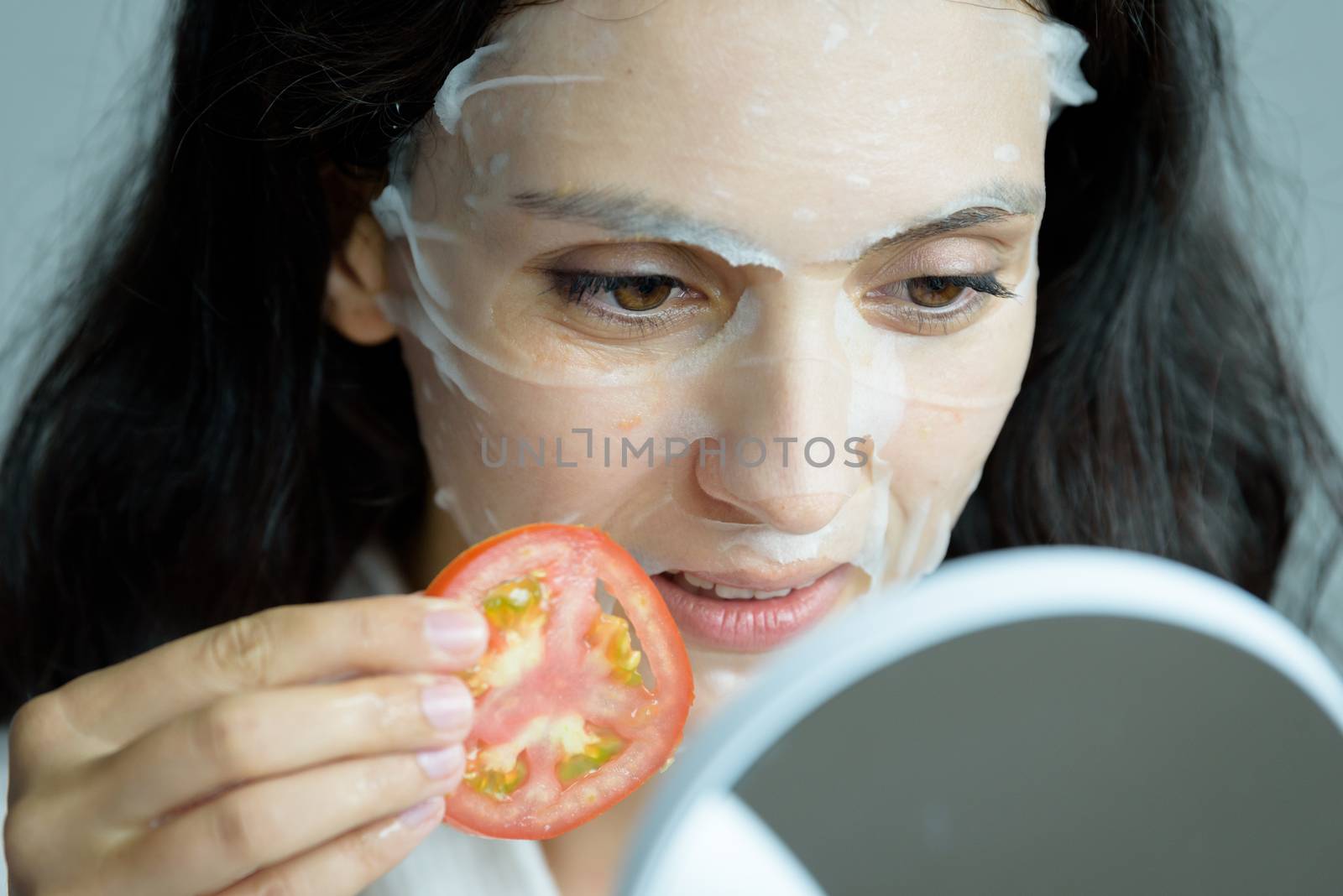 A beautiful woman wearing a towel and a white bathrobe has to look to a mirror for a mask on face-covering eyes with sliced tomatoes with happy and relaxing on the bed at a condominium in the morning.