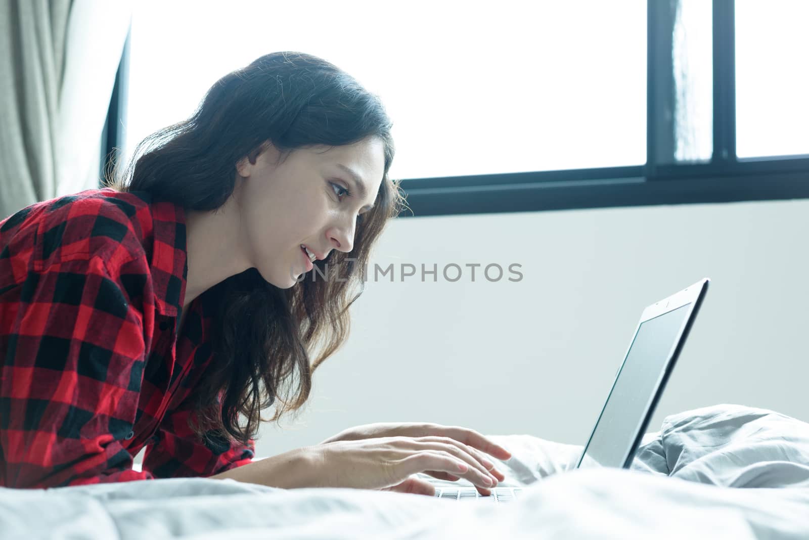 Beautiful woman working on a laptop with smiling and lying down on the bed at a condominium in the morning.