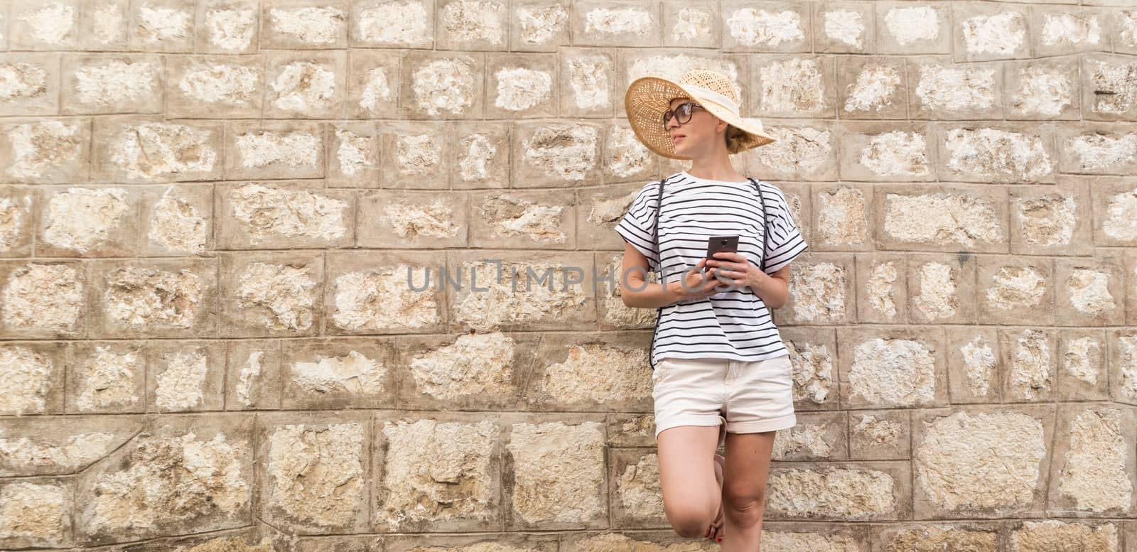 Beautiful young female tourist woman standing in front of old textured stone wall at old Mediterranean town, smiling, holding, smart phone to network on vacationes. Copy space by kasto