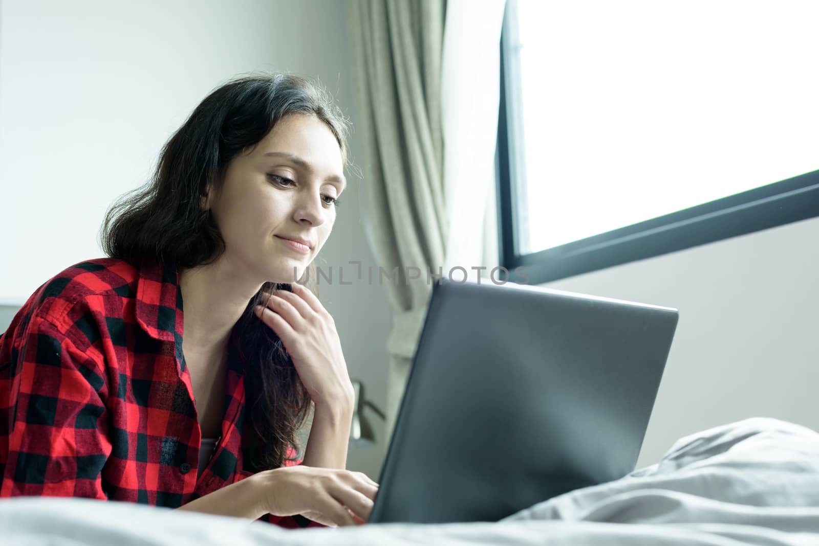 Beautiful woman working on a laptop with smiling and lying down on the bed at a condominium in the morning.