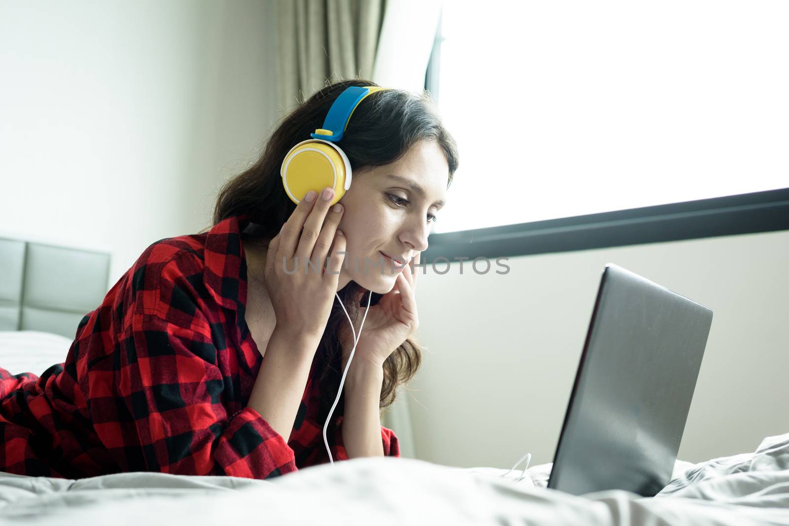 Beautiful woman working and listening to music on a laptop with yellow headphone and lying down on the bed at a condominium in the morning.