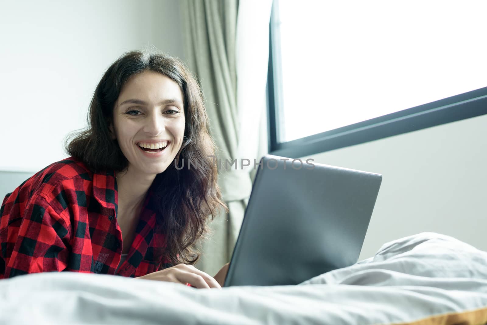 Beautiful woman working on a laptop with smiling and lying down on the bed at a condominium in the morning.