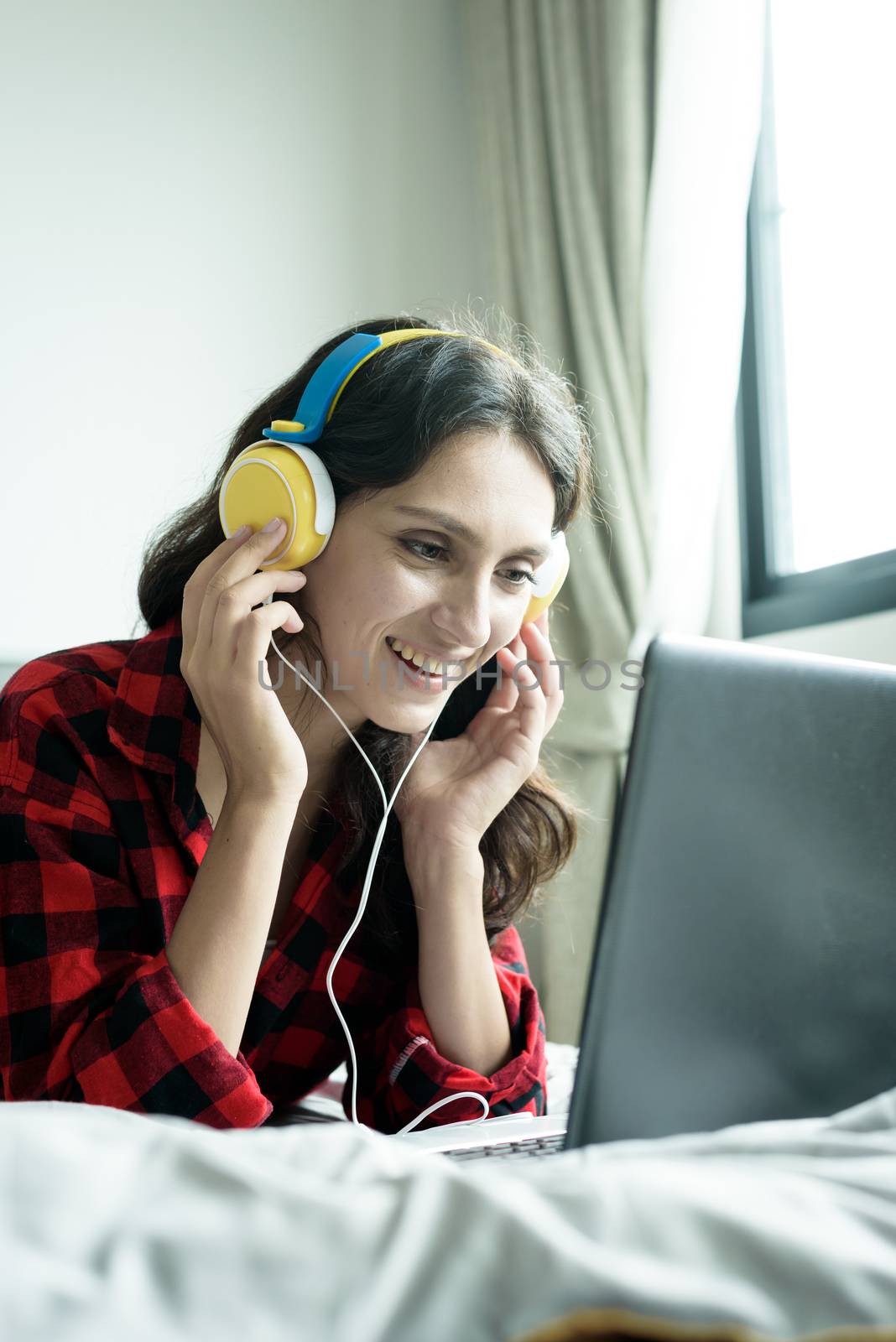 Beautiful woman working and listening to music on a laptop with yellow headphone and lying down on the bed at a condominium in the morning.