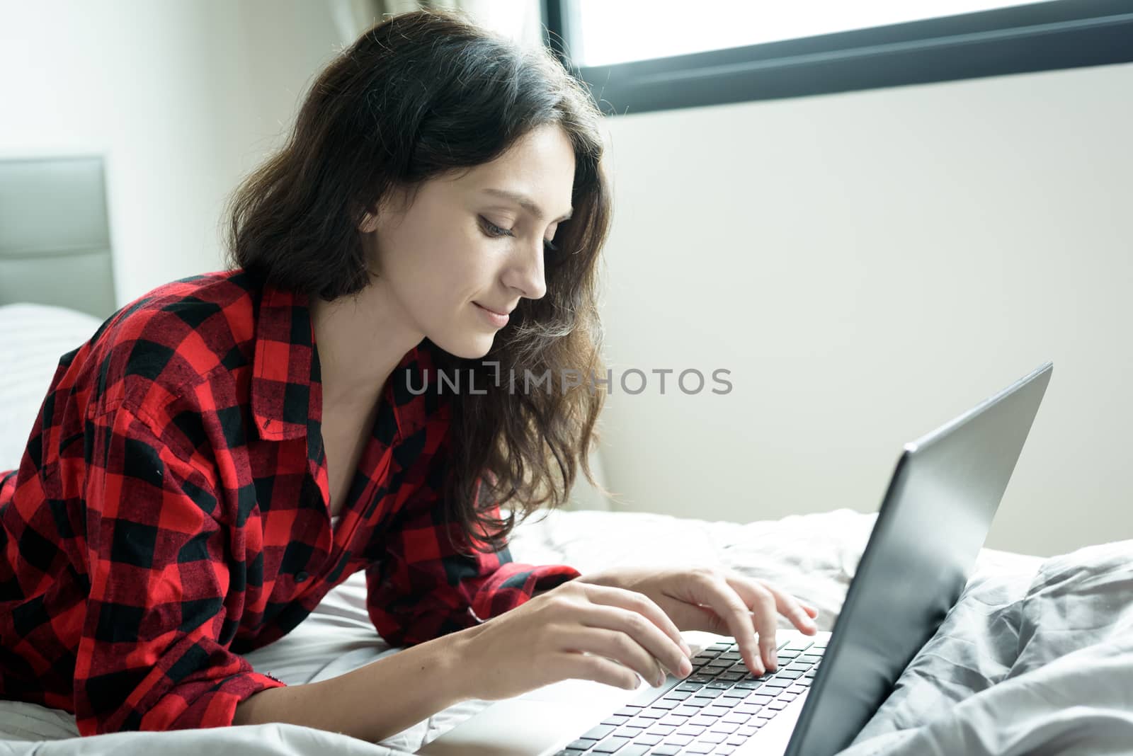 Beautiful woman working on a laptop with smiling and lying down on the bed at a condominium in the morning.