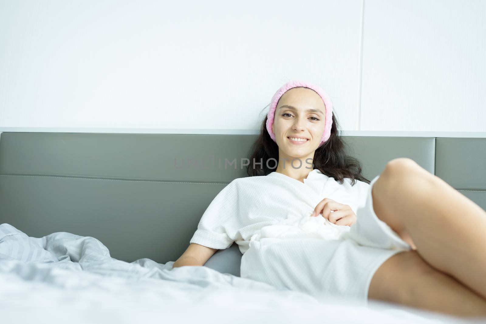 A beautiful woman wearing a towel and a white bathrobe and pink headband with happy and relaxing on the bed at a condominium in the morning.