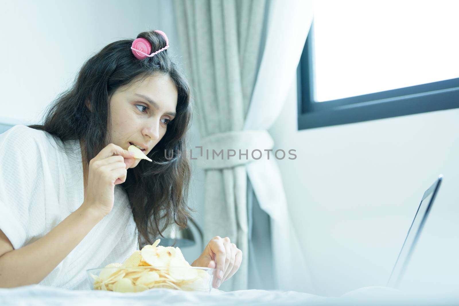 A beautiful woman eating snack potato and playing laptop with lying down on the bed and happiness at a condominium in the morning.