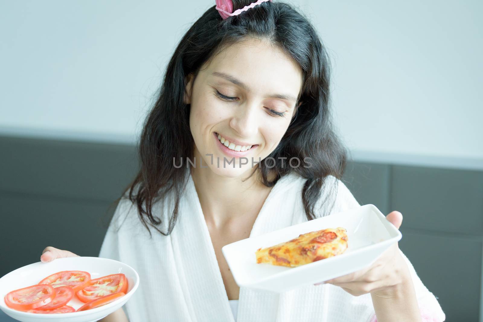 A beautiful woman wearing a towel and a white bathrobe has to eat a pizza and sliced tomato with happy and relaxing on the bed at a condominium in the morning.