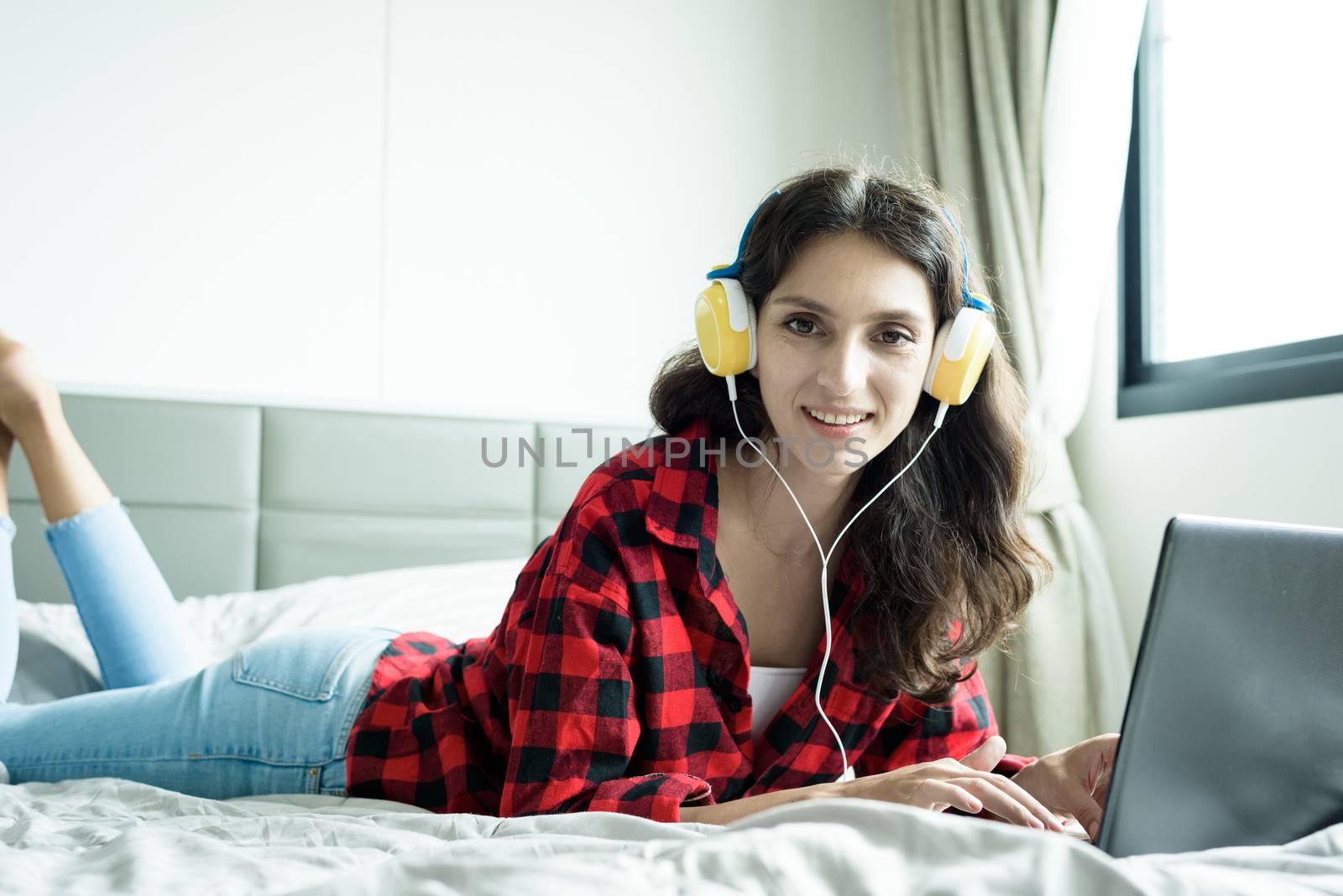 Beautiful woman working and listening to music on a laptop with yellow headphone and lying down on the bed at a condominium in the morning.