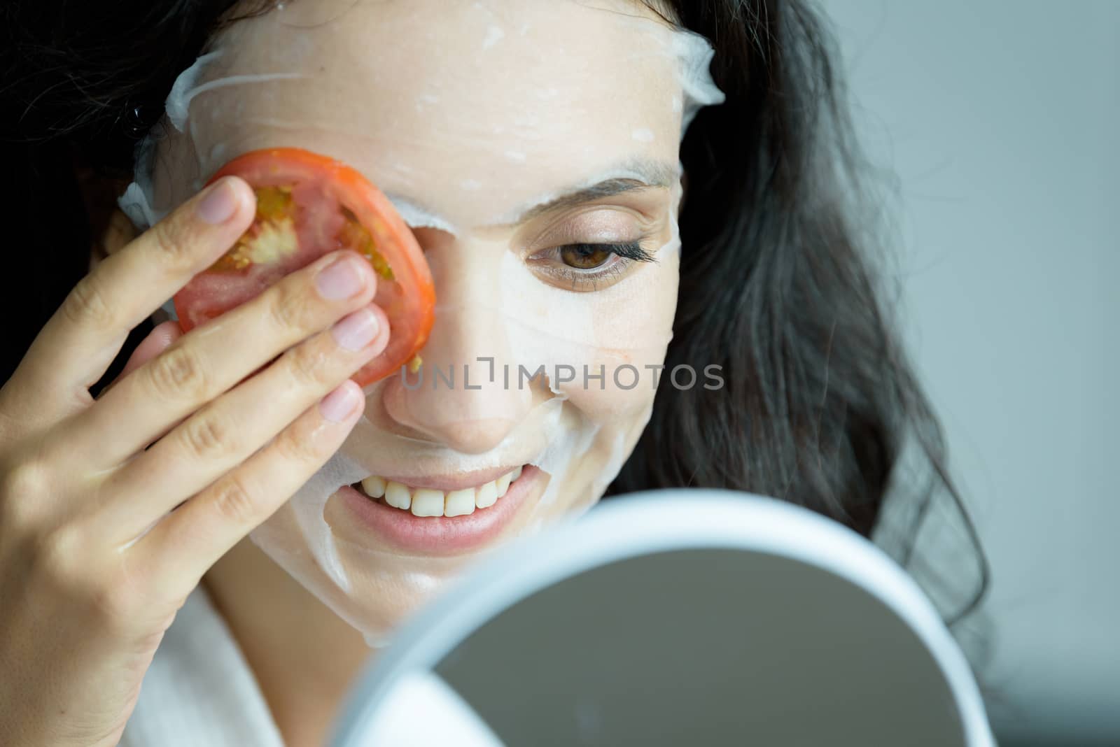 A beautiful woman wearing a towel and a white bathrobe has to look to a mirror for a mask on face-covering eyes with sliced tomatoes with happy and relaxing on the bed at a condominium in the morning.