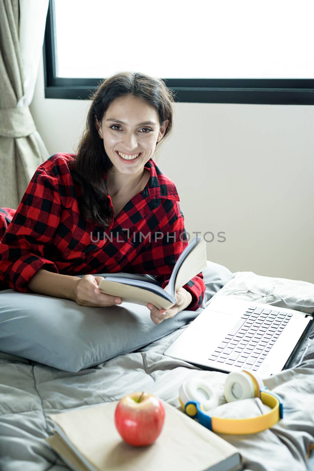 Beautiful woman working and reading a book on a laptop and lying down on the bed at a condominium in the morning.
