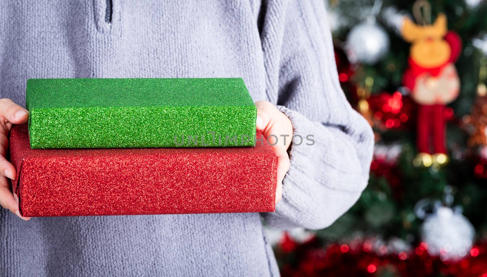 Hands holding a green and red glitter wrapping paper Christmas gift to surprise with Christmas tree background.
