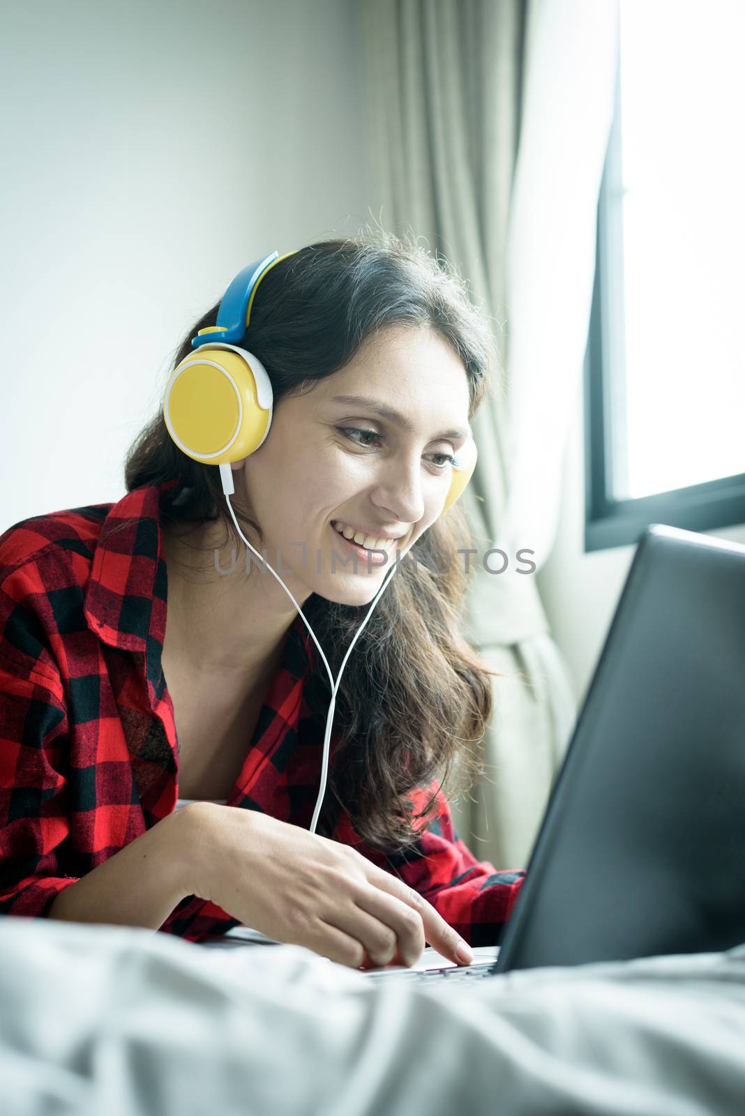 Beautiful woman working and listening to music on a laptop with yellow headphone and lying down on the bed at a condominium in the morning.