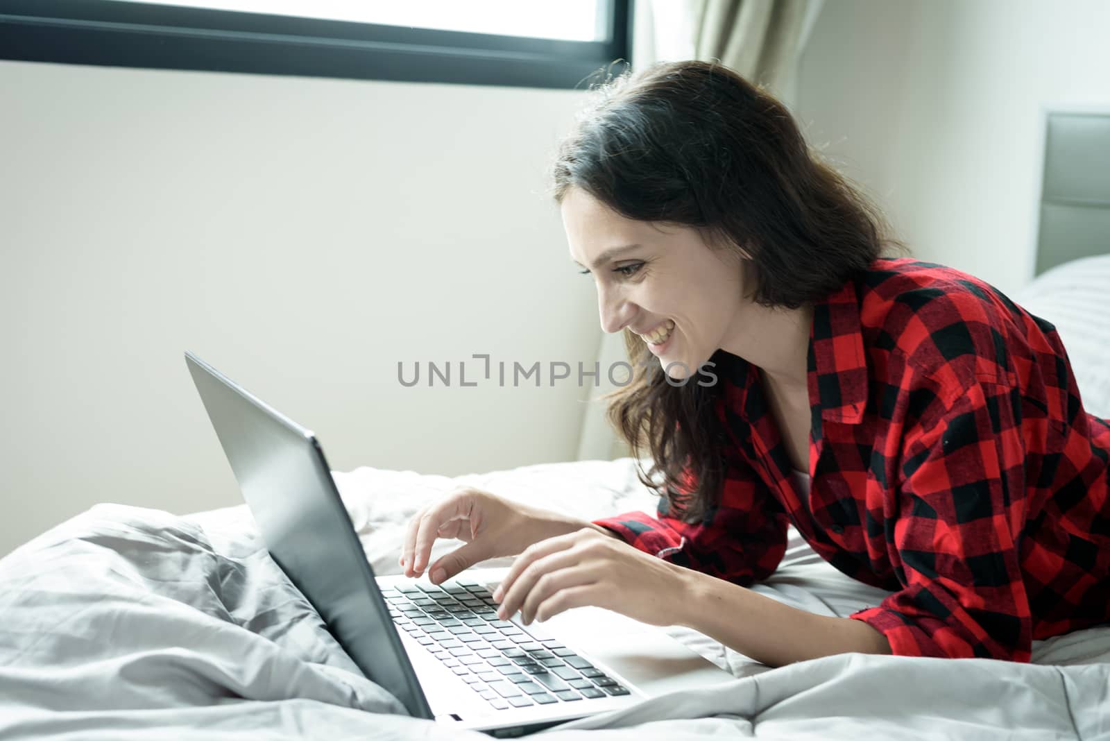 Beautiful woman working on a laptop with smiling and lying down on the bed at a condominium in the morning.