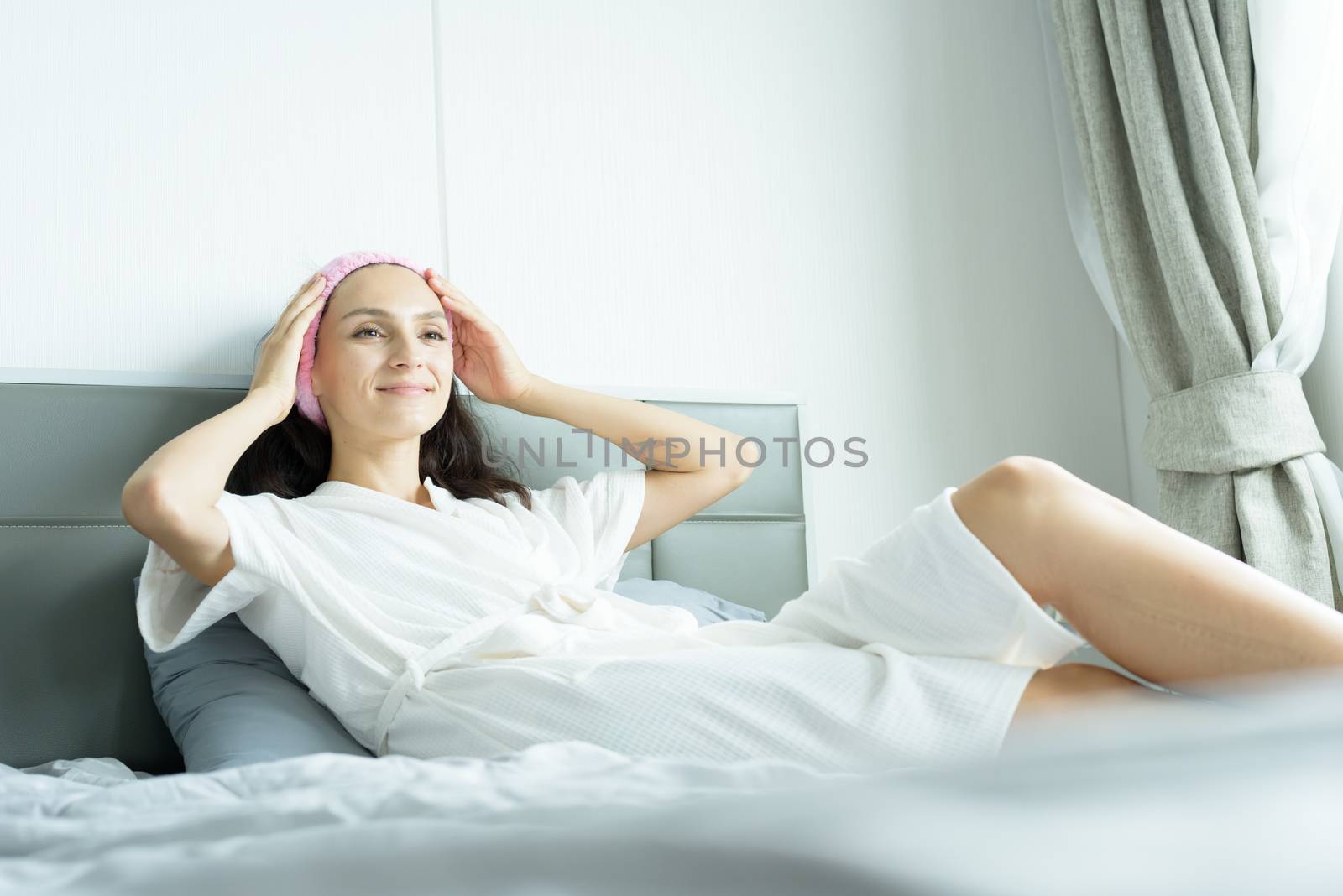 A beautiful woman wearing a towel and a white bathrobe and pink headband with happy and relaxing on the bed at a condominium in the morning.