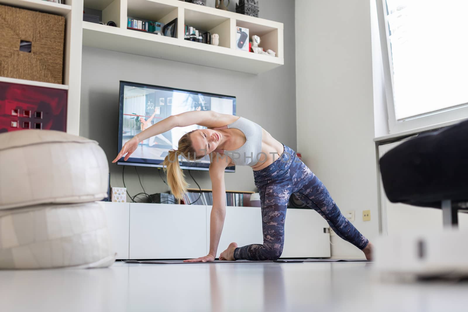 Attractive sporty woman working out at home, doing pilates exercise in front of television in her living room. Social distancing. Stay healthy and stay at home during corona virus pandemic by kasto