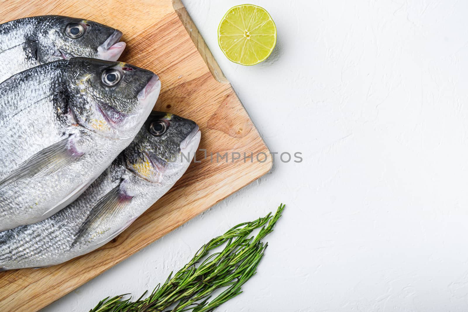 Raw dorada or sea bream fish set on chopping board with herbs for grill uncooked on white textured background top view .