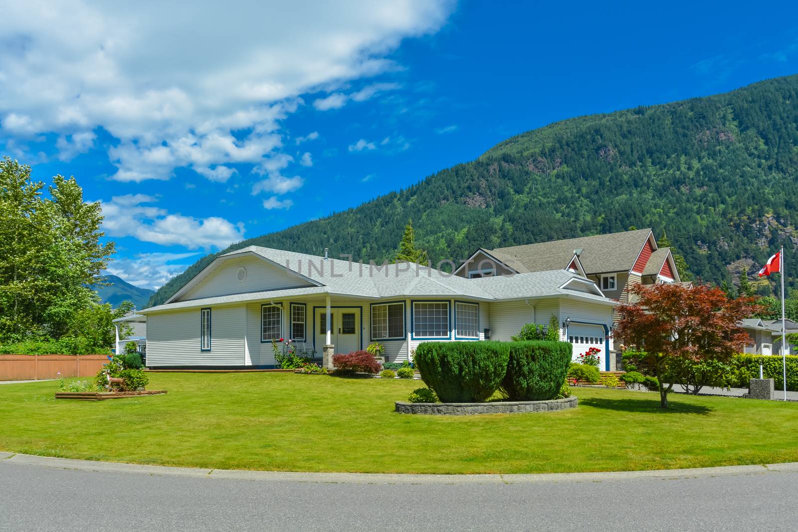 A perfect neighborhood. One level house with Canadian flag on a front yard on blue sky background. Big house with lawn in front and mountain view background. British Columbia, Canada.