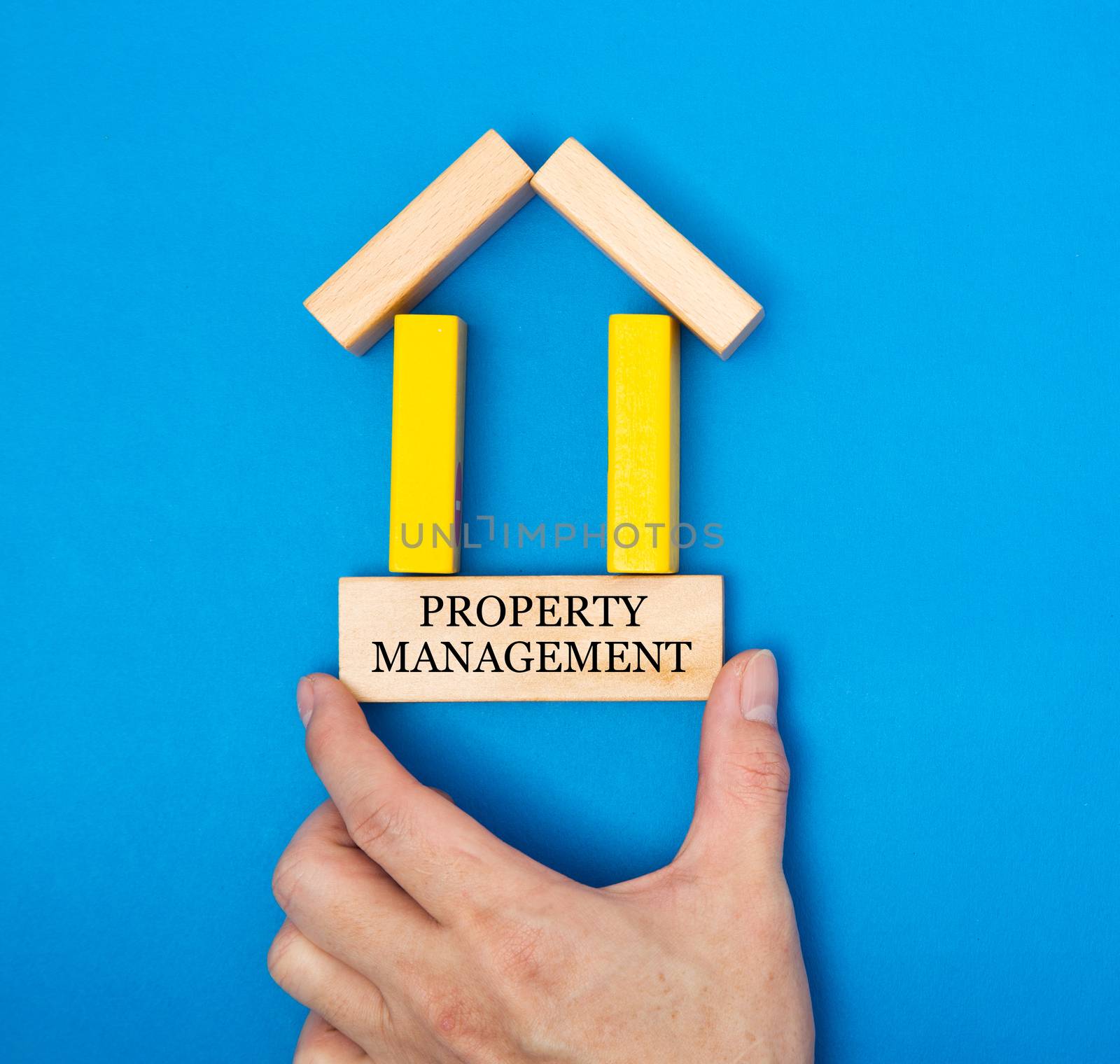 Male hand placing a property management in a house made of wooden blocks in a conceptual image of real estate market. Over blue background.