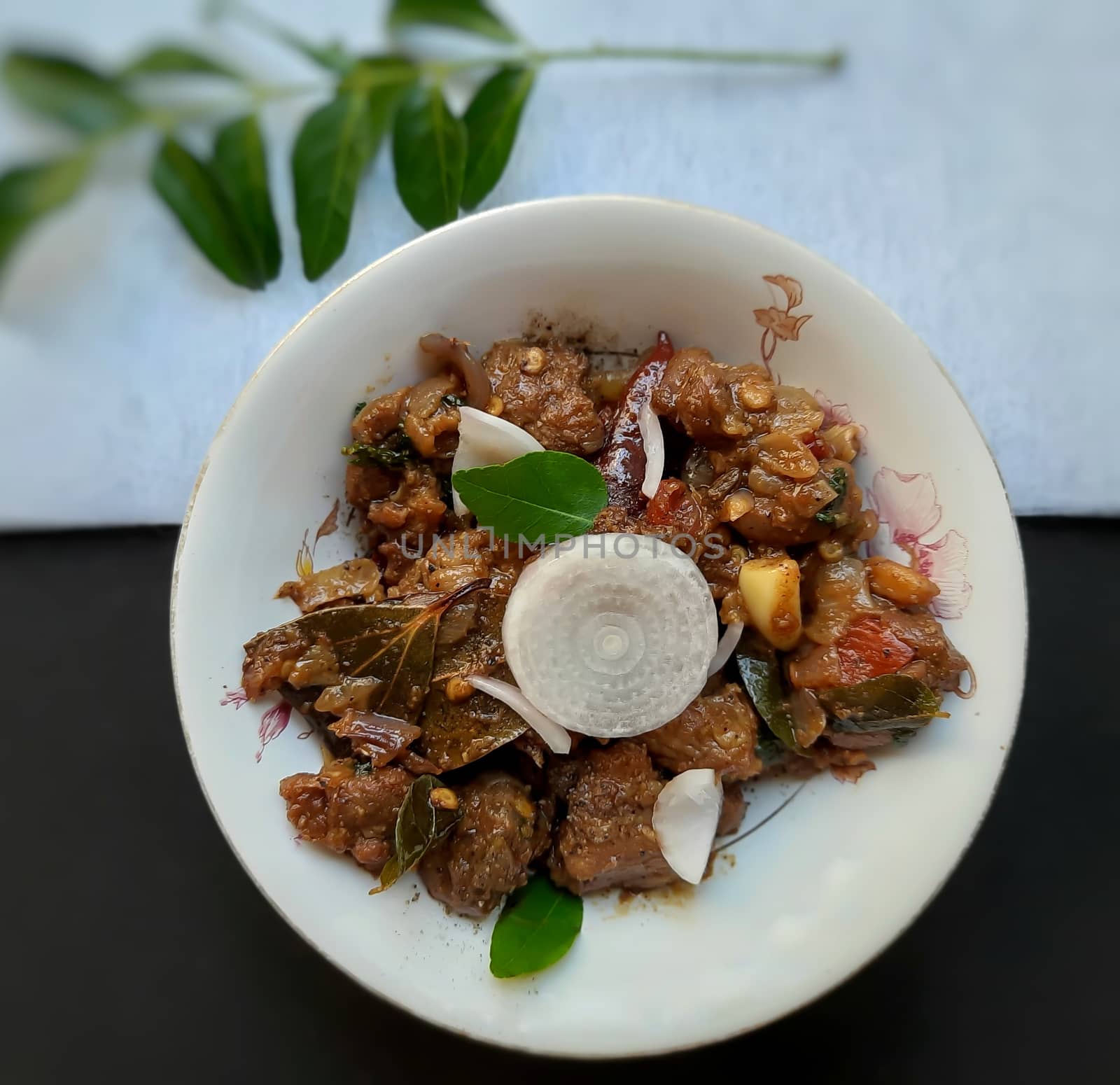 Delicious Indian style spicy pepper mutton placed beautifully in white bowl plated in black and white color paper with onions and curry leaves and good for bones