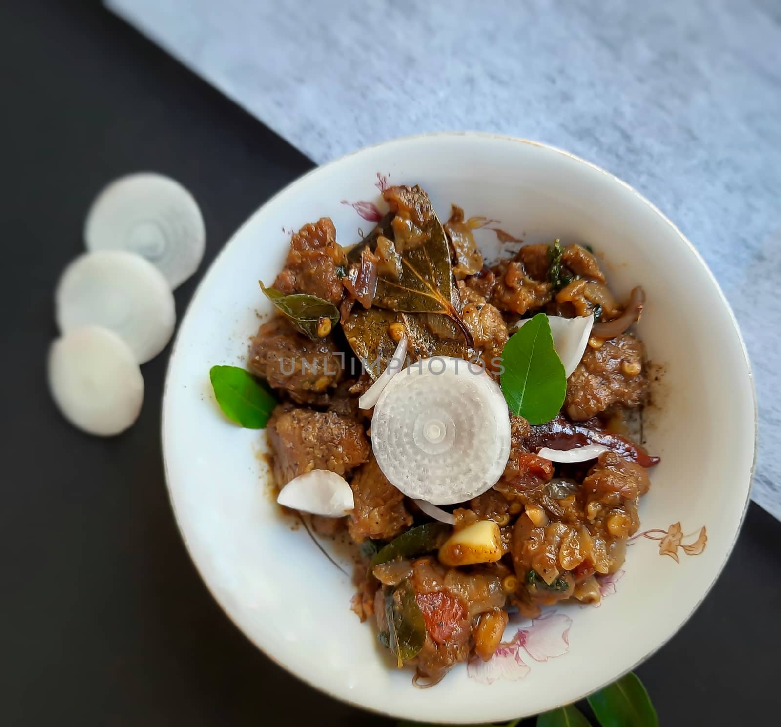 Delicious Indian style spicy pepper mutton placed beautifully in white bowl plated in black and white color paper with onions and curry leaves and good for bones