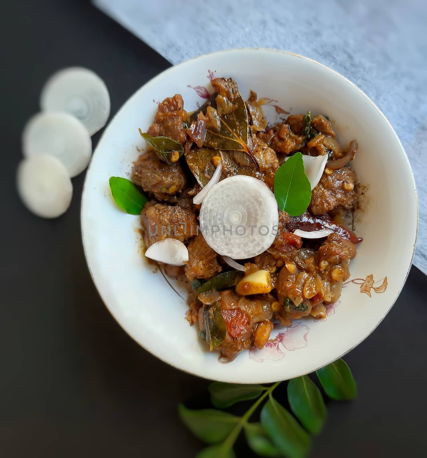 Delicious Indian style spicy pepper mutton placed beautifully in white bowl plated in black and white color paper with onions and curry leaves and good for bones